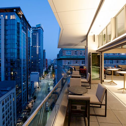 The image shows a modern urban balcony at dusk, with seating and tables. Tall buildings line the lit street below.