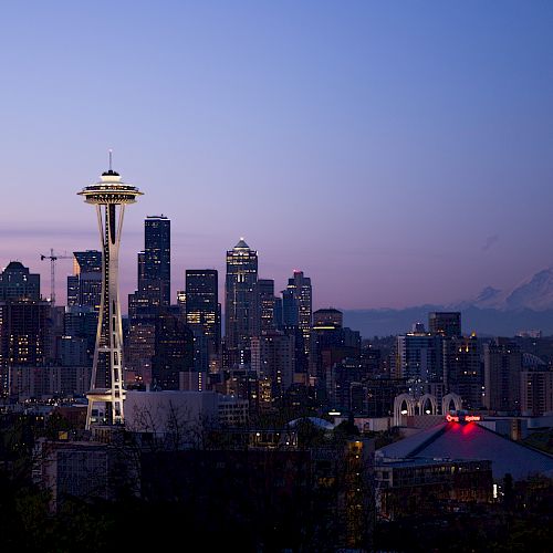 Seattle skyline at dusk with the Space Needle and Mount Rainier visible in the background, against a purple and blue sky.