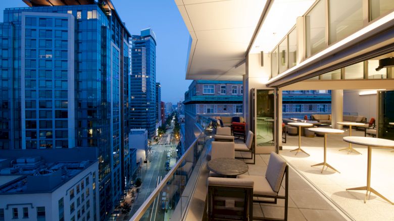 A modern balcony with tables and chairs overlooks a cityscape with tall buildings at twilight.