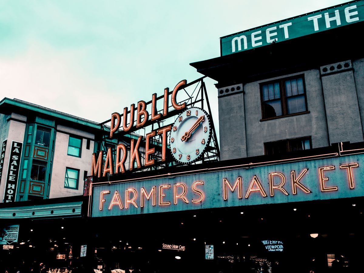 The image shows a vibrant public market sign with a clock above, set against the backdrop of urban buildings in a bustling area.