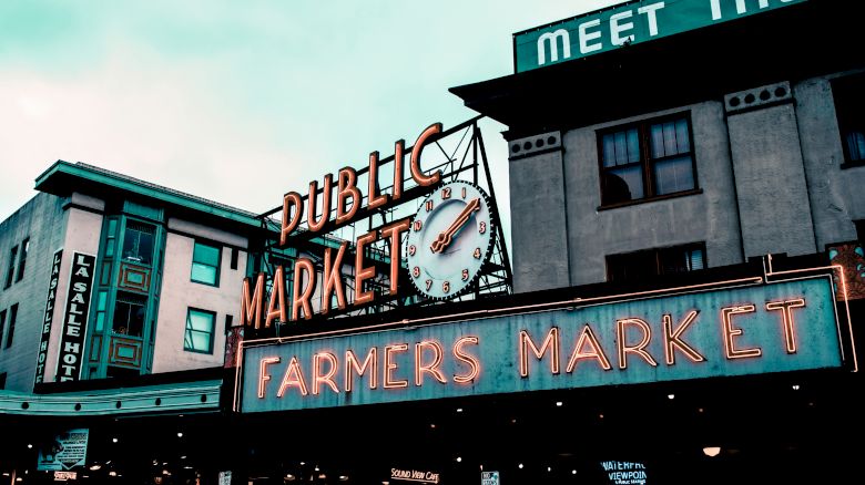 The image shows a vibrant public market sign with a clock above, set against the backdrop of urban buildings in a bustling area.