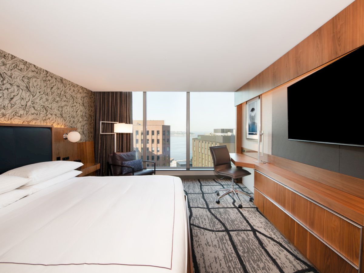 The image shows a modern hotel room with a large bed, a flat-screen TV, a desk with a chair, and a window with a city view.
