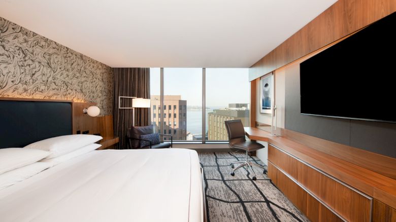 The image shows a modern hotel room with a large bed, a flat-screen TV, a desk with a chair, and a window with a city view.