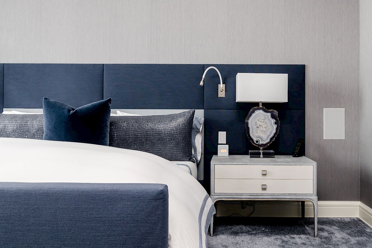 A modern bedroom features a blue upholstered bed, navy and gray pillows, a white nightstand with a lamp, and a decorative item.