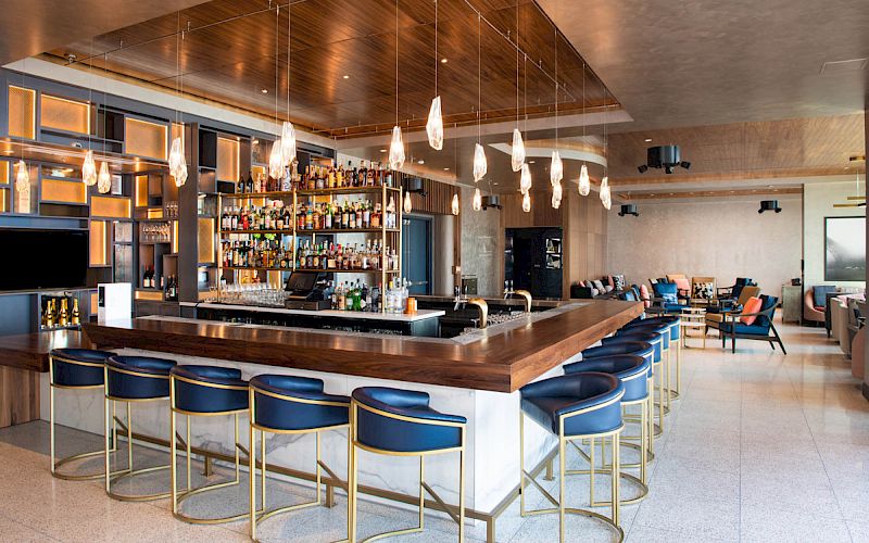 The image shows a modern bar with blue stools, a well-stocked liquor shelf, pendant lights, and a cozy seating area in the background.