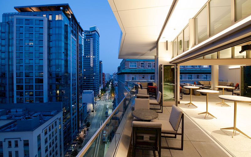 A modern rooftop terrace with seating, overlooking a city street lined with tall glass buildings during dusk.