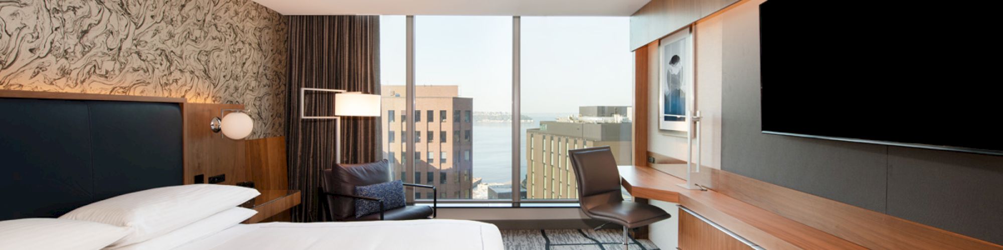 A modern hotel room with a large bed, desk, chair, and TV. Windows offer a view of buildings and water in the background.